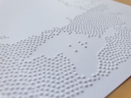 Foto-Nahaufnahme: Ausschnitt aus einem weißen Papier mit vielen erhabenen Punkten, die eine zusammenhängende Fläche bilden. In der Mitte eine unregelmäßig geformte Freifläche mit nur 6 Punkten (die Braille-Buchstaben K und R).
