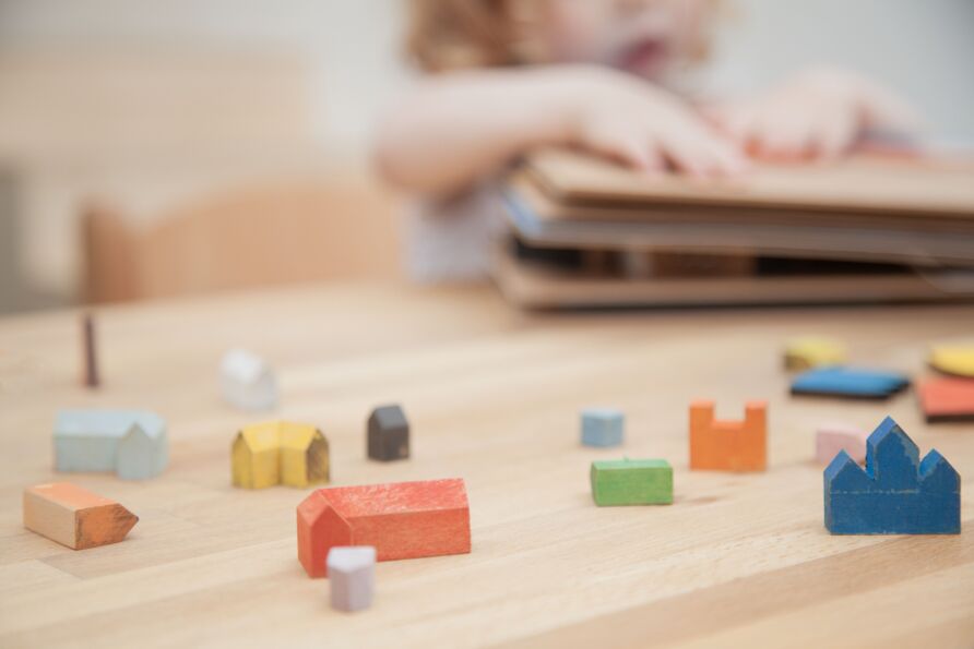 Verschiedene, farbige kleine Häuschen aus Holz auf einem Holztisch. Die Häuser sind auf ihre Grundform reduziert und haben unterschiedliche Formen und Dächer. Im Hintergrund unscharf ein Kleinkind, das die Hände auf ein Tastbuch legt.