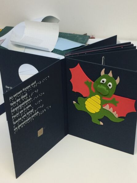 Das Bild zeigt ein aufgeklapptes Tastbuch. Auf der linken Seite steht ein Text in Braille und in Schwarzschrift. Auf der linken Seite ist ein kleiner grüner Drache mit roten Flügeln.