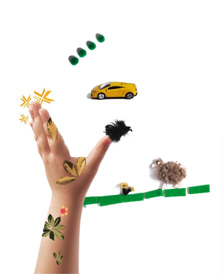 Eine Kinderhand ragt von unten in die Bildmitte. Auf der Haut sind bunte Pflanzenmotive zu sehen, drumherum sind Tastbuchmaterialien arrangiert, darunter ein Spielzeugauto, ein Schaf und ein Vogel.