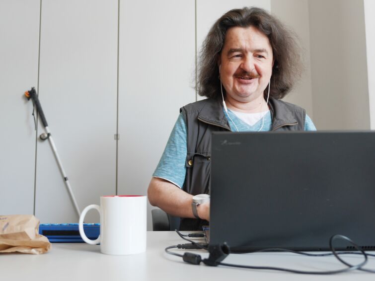 Das Bild zeigt einen braunhaarigen Mann, der vor seinem Laptop sitzt und lächelt. Er hat Kopfhörer im Ohr und hinter ihm sieht man einen weißen Schrank, an dem ein Langstock lehnt.