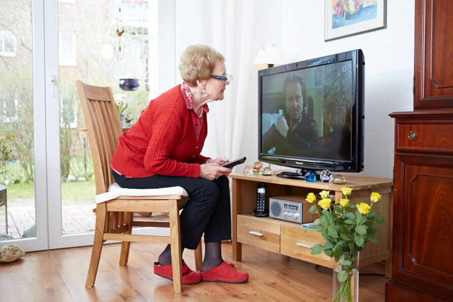 Eine Seniorin mit blonder Kurzhaarfrisur sitzt in einer Wohnung vornübergebeugt auf einem Stuhl. Sie betrachtet aus 30 cm Abstand durch eine Vergrößerungsbrille einen Flachbildfernseher, zu sehen ist ein Paar.