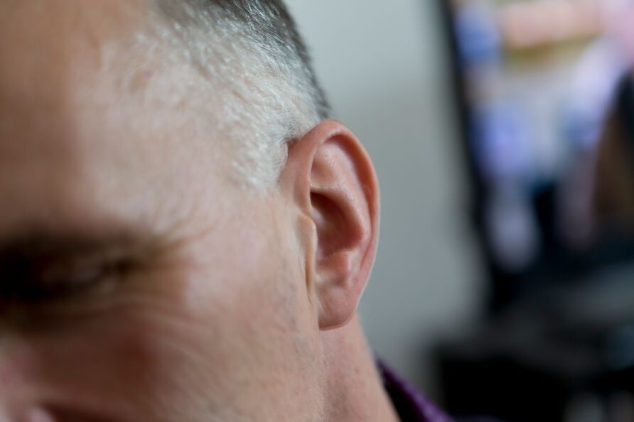 Ein menschliches Ohr in Großaufnahme, im Hintergrund ein unscharfes Fernsehbild