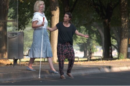 Eine Frau mit entschlossenem Gesichtsausdruck greift einer sehbehinderten Frau mit Langstock an den Arm und möchte sie über eine Straße führen. Beide tragen sommerliche Kleidung.