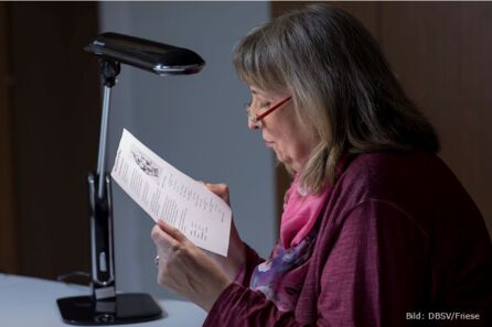 Foto: Eine Frau mittleren Alters mit schulterlangen, grauen Haaren liest ein bedrucktes Blatt Papier, das sie unter eine Leseleuchte hält. Sie trägt Oberbekleidung in Dunkelviolett und ein dazu passendes Halstuch.