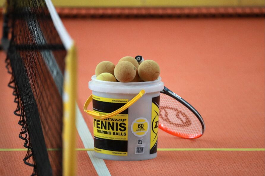 Ein Eimer voller Tennisbälle steht auf dem Tennisplatz unmittelbar vor dem Netz. Ein Tennisschläger wurde an den Eimer gelehnt.