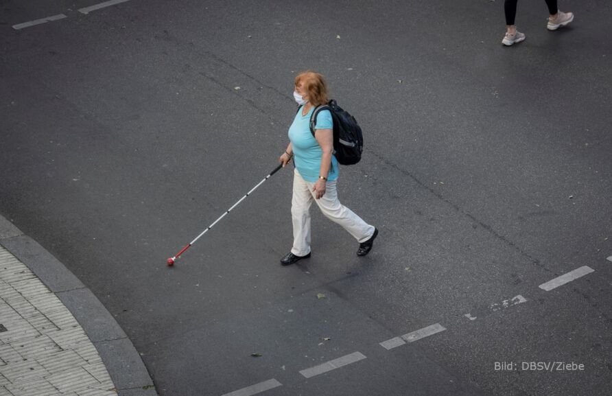 Foto: Blick aus erhöhter Position auf eine Seniorin mit Langstock und Mund-Nasen-Bedeckung, die eine Straße kreuzt. Sie hat rotblonde Haare, trägt eine naturweiße Hose, ein blaues T-Shirt und einen dunklen Rucksack.