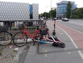 An einem Metallgeländer lehnen Fahrräder, daneben stehen und liegen E-Roller mitten im Weg.