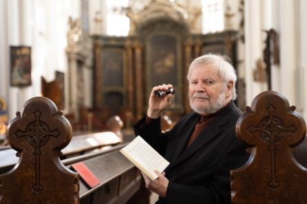 Foto: Ein älterer Herr mit weißem Haar und Bart sitzt in einer Kirchenbank und schaut den Betrachter an, in der linken Hand ein aufgeschlagenes Gesangbuch, in der rechten ein Monokular.