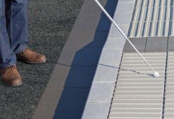 Die abgesenkte Bordsteinkante eines Gehwegs, sie ist farblich abgesetzt. Auf dem Gehweg befinden sich taktile Bodenindikatoren mit Rillen. Eine Person, von der nur die Beine zu sehen sind, steht vor dem Gehweg. Ihr Langstock berührt eine Bodenplatte.