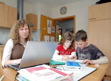 An einem Küchentisch sitzt eine Frau konzentriert vor einem Laptop. Zwei Kinder sitzen mit ihr am Tisch und lesen gemeinsam in einem Buch. Der Rest des Tisches ist von Lehrbüchern bedeckt.