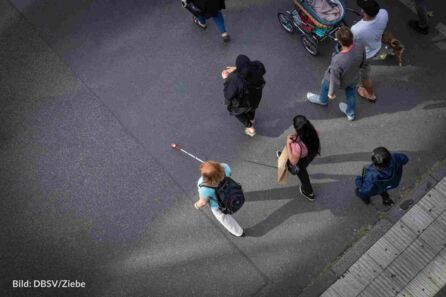 Foto aus der Vogelperspektive: Eine Frau mit Langstock läuft neben anderen Passanten über eine Straße. Sie hat rotblonde Haare, trägt eine weiße Hose, ein hellblaues T-Shirt und einen dunklen Rucksack.