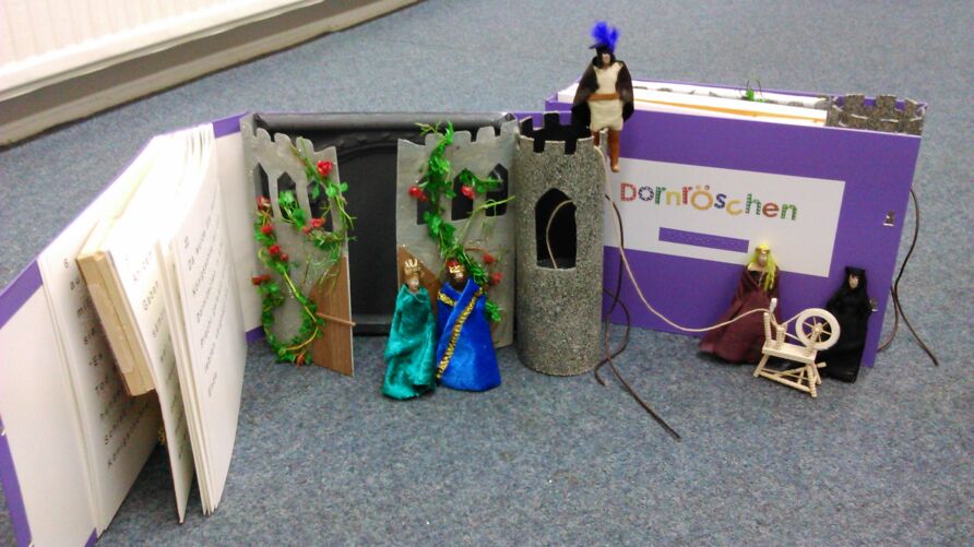 Verschiedene Elemente des Tastbuches Dornröschen. Das Königspaar steht vor dem Schloss mit der Rosenhecke. Der Prinz steht oben auf dem Turm. Dornröschen und die böse weise Frau stehen mit dem Spinnrad vor der Titelseite des Buches.