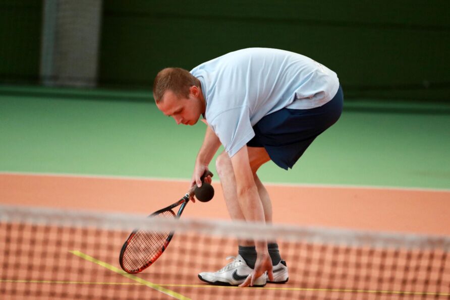 Auf dem Tennisplatz steht ein Mann in Sportkleidung (helles T-Shirt, kurze Hose, Sportschuhe). In der rechten Hand hält er den Tennisschläger und einen Ball. Er beugt sich auf den Boden und berührt mit der linken Hand eine Markierungslinie.