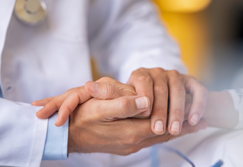 Eine Hand wird von zwei anderen Händen gehalten. Die Hände gehören zu einer Person, die unscharf im Hintergrund zu erkennen ist. Sie trägt einen weißen Arztkittel und um den Hals ein Stethoskop.