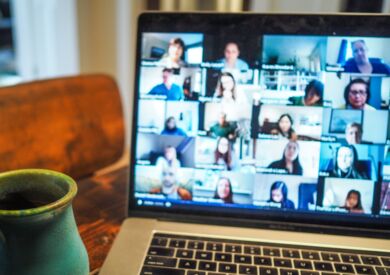 Ein aufgeklappter Laptop, auf dem Bildschirm sind Kacheln angeordnet, die die Teilnehmenden einer Videokonferenz zeigen. Am linken Rand ragt eine Keramiktasse ins Bild hinein.