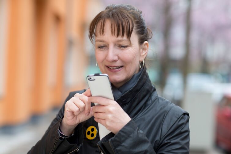 Auf dem Bürgersteig: Eine Frau mittleren Alters hält ein Smartphone in der Hand und tippt mit dem Finger ihrer rechten Hand aufs Display. Sie lächelt. An ihrer schwarzen Jacke ein gelber Button mit drei schwarzen Punkten..