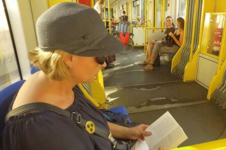 Eine Frau mit Brille und schwarz-gelbem Blindenabzeichen auf dem Handtaschenriemen sitzt in der Straßenbahn und liest in einem kleinen Heft. Weiter hinten sitzen schräg gegenüber zwei weitere Frauen. Eine von ihnen schaut den sehbehinderten Fahrgast an.