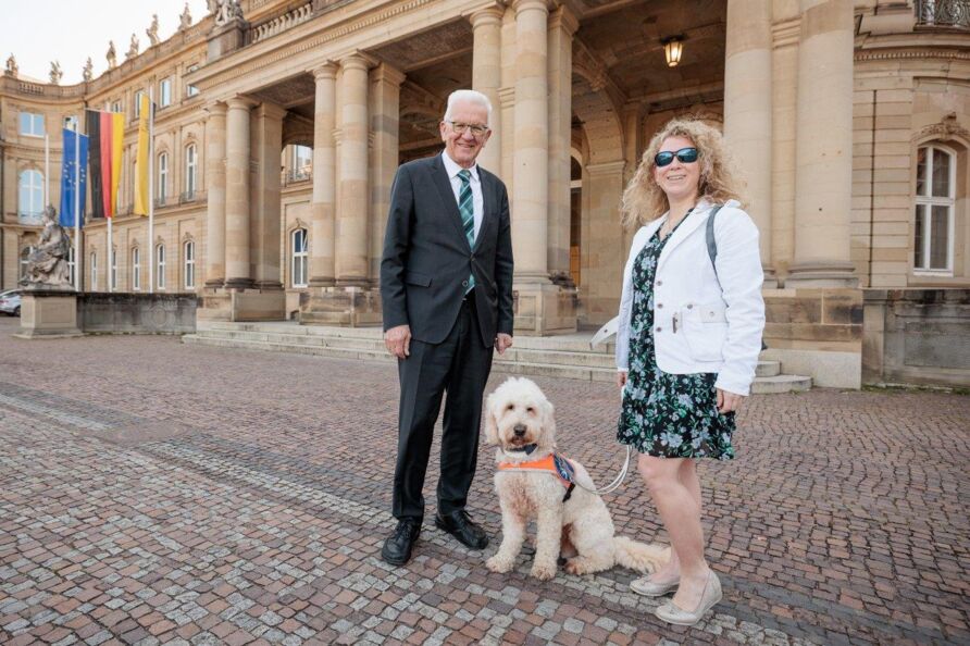 Vor dem Stuttgarter Neuen Schloss steht links Winfried Kretschmann mit weißem Haar, Brille und schwarzem Anzug. Rechts steht Lisa Mümmler, eine junge, blonde Frau mit Sonnenbrille im geblümten Kleid. In der Mitte ihr Hund Harry mit hellem lockigem Fell.