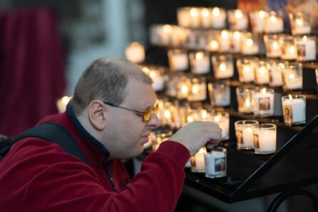 Foto: Schulter- und Kopfpartie eines jungen Mannes im Profil. Er trägt eine Brille mit dicken Gläsern und zündet aus nächster Nähe eine Kerze an. Im Hintergrund weitere leuchtende Kerzen.