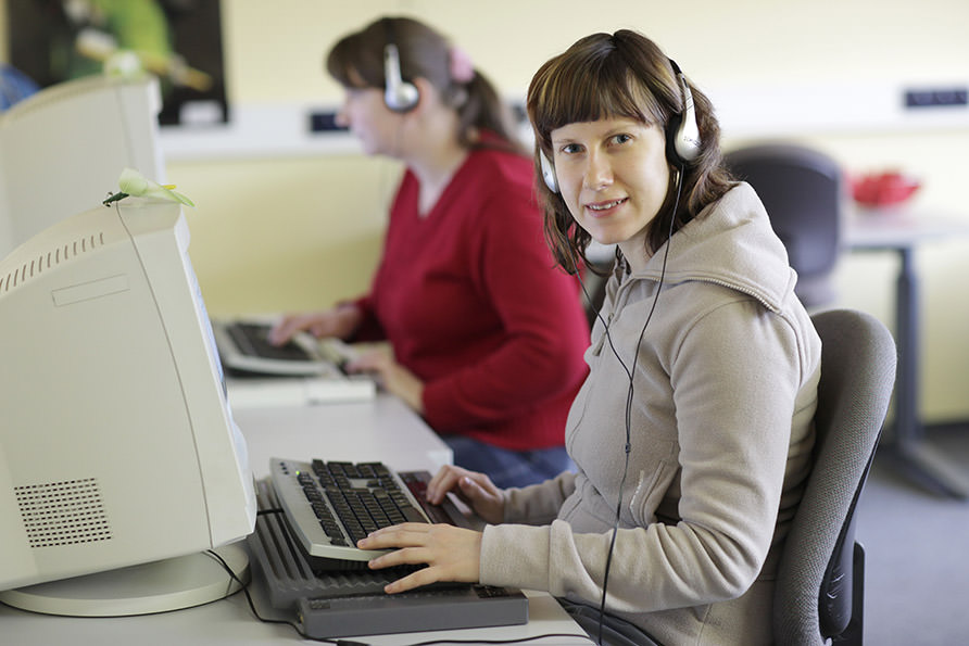Zwei Damen arbeiten an einem Computer. Beide nutzen eine Braille-Zeile an der Tastatur und tragen Kopfhörer. Die vordere Dame blickt in Richtung des Betrachters.