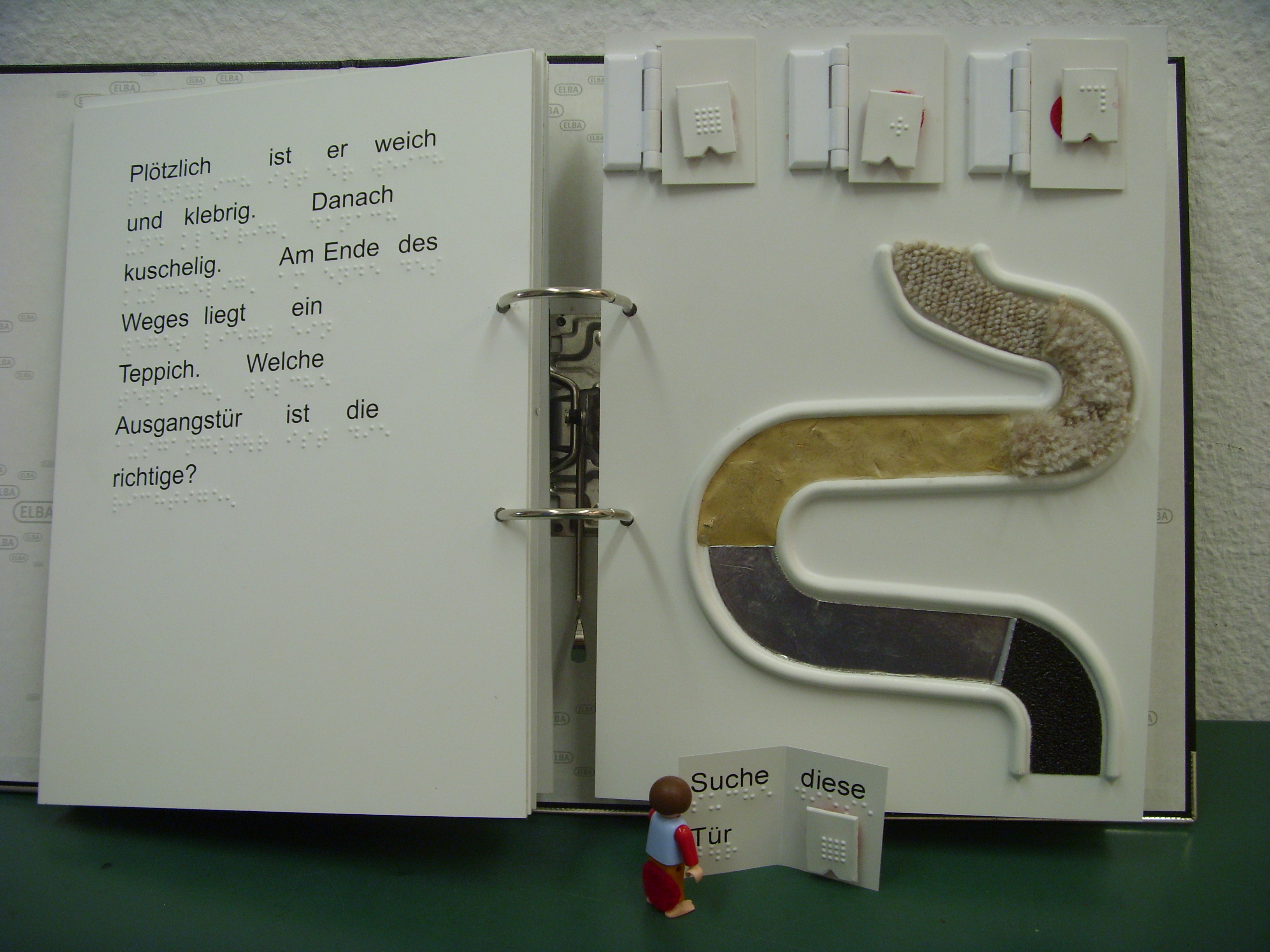 Doppelseite: links, Text; rechts, ein Weg mit Kurven aus unterschiedlichen Materialien, drei Türen mit Punktschrift-Kärthchen und eine Playmobil-Figur